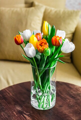 Bouquet of colorful tulips in a glass vase on a wooden table against the background of a yellow sofa and pillows. Home still life