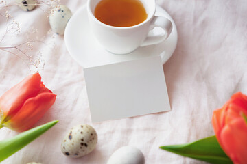 blank paper business card on light fabric with cup of tea, , quail eggs and tulips. Easter mock up