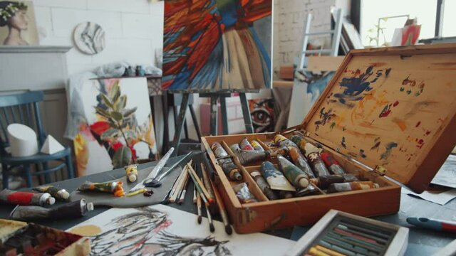 Zoom out shot of wooden case with oil paint tubes and different art supplies for painting and drawing on worktable in studio