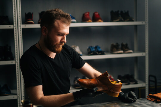Portrait of handsome bearded shoemaker examining light brown leather shoes during restoration working. Concept of cobbler artisan repairing and restoration work in shoe repair shop.