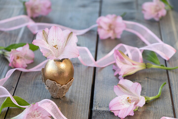 Pink alstroemeria flower in a golden egg on a wooden background with pink flowers and pink ribbon. Happy Easter concept.