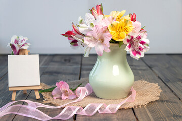 Bouquet from pink, yellow, magenta, white and red alstroemerias in pastel green vase with pink ribbon near small empty easel on wooden background.Happy birthday or Holiday concept.