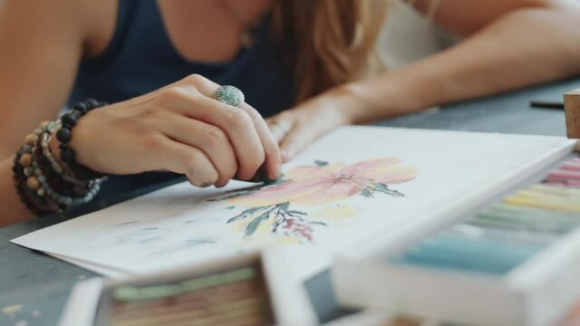 Zoom in tilt up shot of young Caucasian woman drawing beautiful pastel flower on paper with crayons at desk in art studio