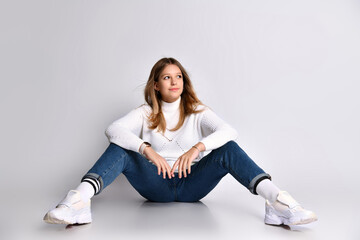 Cute teenage girl with long hair, smartwatch. Dressed in jeans, a sweater and sneakers. She is sitting on the floor and smiling, isolated on white.