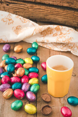 Cup of espresso and colorful wrapped easter chocolate eggs on a rustic wooden table, vertical