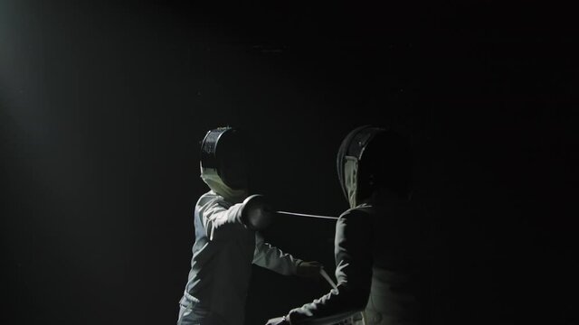 Two professional fencers demonstrate their mastery of foil fencing. Silhouettes of athletes in white uniforms and protective masks fighting duel on black studio background. Slow motion. Close up.