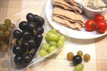 Assorted grapes in plastic container with a plate of humous,  pitta bread and tomato
