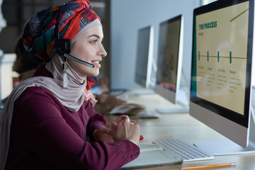 Young muslim woman in headphones looking at computer monitor and talking on the phone working in...