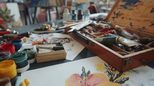 Zoom in shot of metal tubes with oil paint and brushes in wooden case surrounded by art supplies on worktable in studio