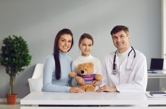 Smiling Mother, Child And Family Practitioner Looking At Camera Sitting At Table At Doctor's Office All Together. Portrait Of Happy Mom, Healthy Kid And Friendly Pediatrician In Modern Health Center