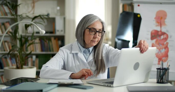 Woman in glasses and gown opening laptop and started typing while entering information from form.Elderly female doctor working with personal computer and documents in medical office.