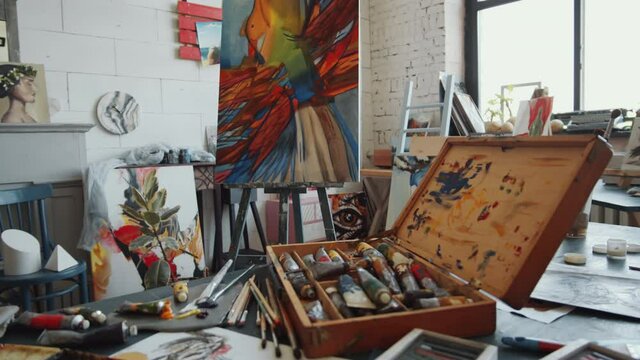 Zoom in shot of lots of art supplies on worktable and beautiful abstract painting on easel in studio