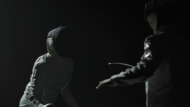 Two professional fencers demonstrate their mastery of foil fencing. Silhouettes of athletes in white uniforms and protective masks fighting duel on black studio background. Slow motion. Close up.