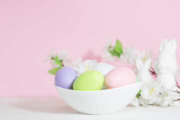 Easter eggs, pink, green and lilac, in a white plate, on a concrete white table, next to bunnies and flowers, on a pink background. Pastel shades.
