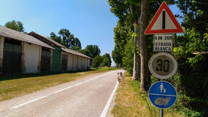 Asphalt road in the middle of the Veneto countryside, Italy. Sheds covered with asbestos, trees along the road, a bicycle on the edge, four road signs on a pole. Written in Italian: a m 200 white road