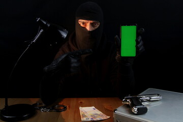 Criminal man wearing balaclava holding smartphone with green screen pointing with finger, over...