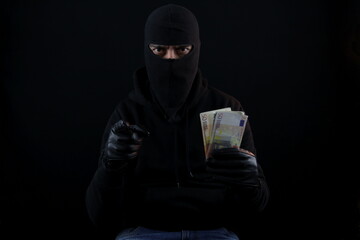 Man wearing balaclava sitting, holding banknotes and pointing finger at the camera. Crime concept