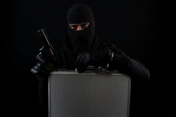 Man with balaclava sitting, holding gun and briefcase attached wrist with handcuffs. Crime concept