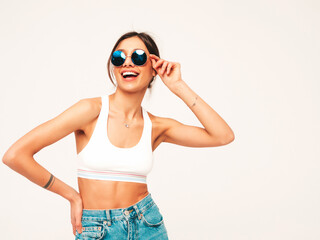Obraz premium Beautiful smiling woman dressed in white jersey top shirt and jeans. Sexy carefree cheerful model having fun indoors.Adorable and positive female posing on grey background in studio in sunglasses
