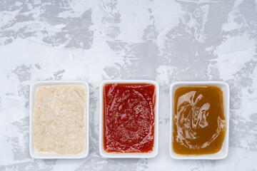 Three kinds of sauces on a white cement background: traditional classic ketchup, white sauce, sweet and sour Chinese sauce. Top view copy space