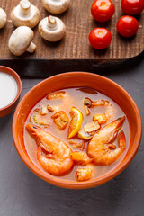 Tom yam soup with shrimps in a plate on a wooden table on a napkin next to a bowl with coconut milk mushrooms and tomatoes.