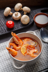 Tom yam soup with shrimps in a tureen on a concrete background next to a bowl with coconut milk tomatoes and mushrooms on a wooden stand.