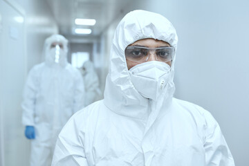 Portrait of male doctor in protective uniform and mask looking at camera while standing in the corridor and working during pandemic