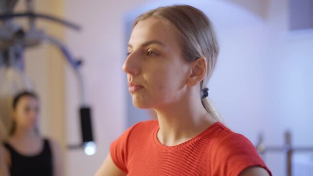 Headshot of positive confident Caucasian sportswoman stretching neck muscles in gym with blurred sportswoman resting at background on exercise machine. Smiling slim athlete working out indoors.