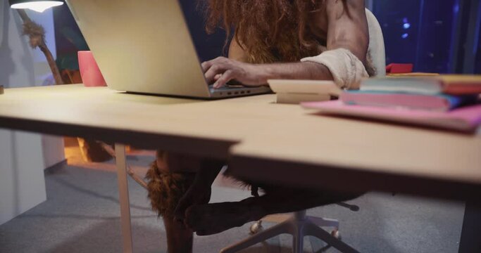 Full-length shot of nomadic prehistoric human homo sapiens brutal bearded neanderthal wearing animal coat using laptop computer in office.