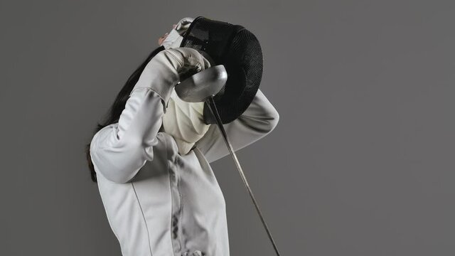 Portrait of young woman fencer with rapier in her hands puts on and then removes protective mask on gray studio background. Sportswoman posing for fencing sports school poster. Slow motion. Close up.