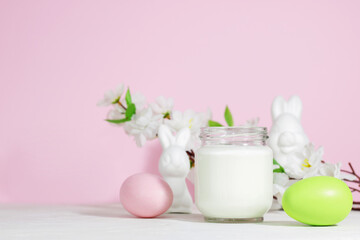 Obraz na płótnie Canvas Organic probiotic milk kefir drink or yogurt in a glass container, with colored Easter eggs, on a pink background with a rabbit. Easter concept. Cold fermented milk drink with probiotics.