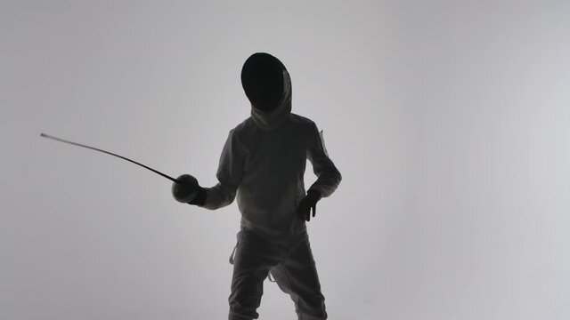 Silhouette of young boy in white uniform and mask masterfully wields rapier on gray studio background. Practicing attacking and defense skills, fencing lesson at sports school. Slow motion. Close up.