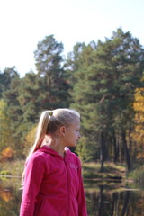 A teenage girl with long blond hair stands on the shore of a lake against the backdrop of water and autumn yellow forest in autumn sunlight. Portrait of a blond girl in profile.