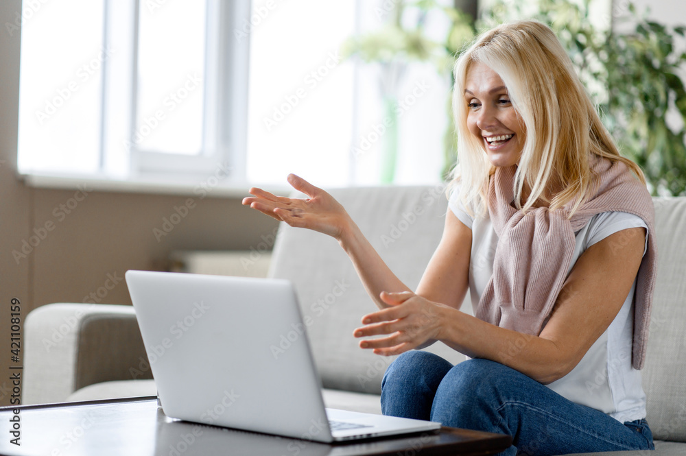 Wall mural A beautiful blonde is sitting at a laptop in the living room on the couch. A middle-aged caucasian happy woman communicates via video call uses laptop with friends or family, telecommunication concept