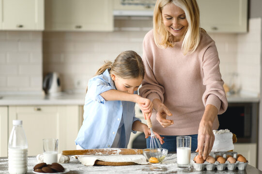A middle-aged caucasian woman teaches a little granddaughter to cook. Grandma teaches her granddaughter to beat eggs, they make and roll the dough, use ingredients, cooking together