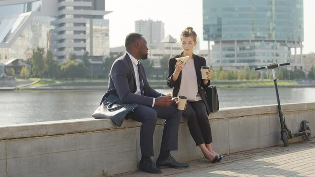 Full shot of cheerful multiethnic couple sitting at river embankment n city centre having lunch together. Diverse business-like couple chatting while eating sandwiches and drinking coffee outdoors