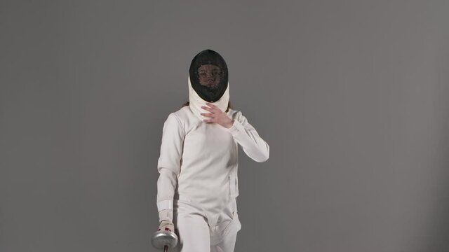 Portrait of charming female fencing athlete in white uniform on gray studio background. Young woman removes mask and poses with foil in hands for sports fencing school poster. Slow motion. Close up.