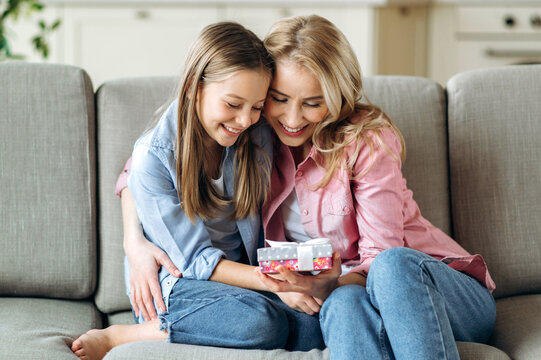 Mom And Daughter Hugging Each Other. A Blond Caucasian Mom And Her Preschooler Girl Sitting On The Sofa In The Living Room And Looks At The Gift Box, Beautifully Decorated With A Bow, Smiling