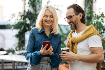 Two caucasian adult friends or coworkers are watching something on smartphone. A middle-aged caucasian man and a beautiful blond woman browsing internet or social media while coffee break