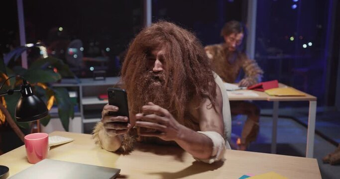 Nomadic neanderthal caveman in animal fur sitting in corporate modern office using mobile phone interacting internet taking selfie having fun. Evolution process.
