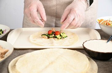 A woman in transparent gloves lays chopped vegetables, cucumbers and tomatoes on a round pita bread.