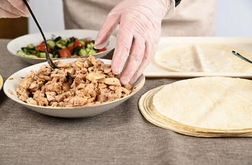 A woman places chopped fried chicken on a round pita bread by a tablespoon.