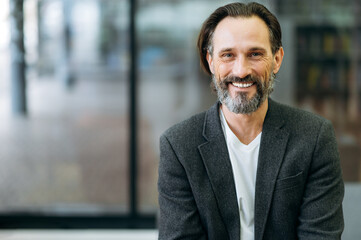 Obraz premium Portrait of a handsome office worker or ceo. Confident stylish bearded middle aged caucasian man in formal jacket and white t-shirt smiles and looks directly into the camera