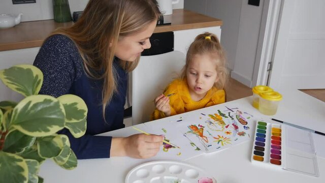 Mother and daughter painting at home. Cute little kid in yellow sweater having fun with parent and paints. Concept of early childhood education, hobby, talent, preschool leisure and parenting