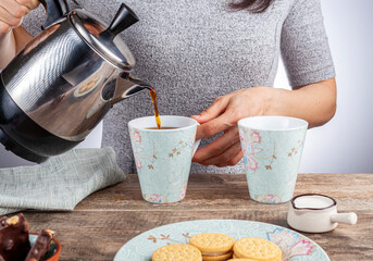 a tea time or coffee time concept with sandwich cookies, bars of chocolate aesthetic ceramic mug and plates as well as a mini creamer pitcher on wooden table. A woman is pouring hot drink into mug.