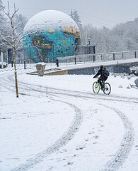 Duringa snow storm, a man on bicycle in riverfront park with the eco-earth globe map in background,...