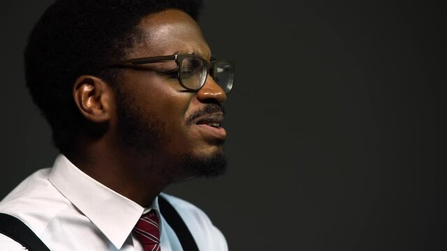 Handsome African American man in a stylish suit and glasses sings with pleasure, closing his eyes. A black performer performs on a gray background in the studio. Close up.