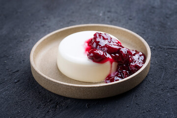 Modern style traditional blancmange almond pudding sour cherry coulis served as close-up in a Nordic design plate on black background