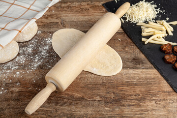 Closeup image A baker's wooden rolling pin on  a flattened dough on wood kitchen countertop. A bread making concept image with loafs and cheese and sucuk in back. Turkish kasarli sucuklu pide recipe