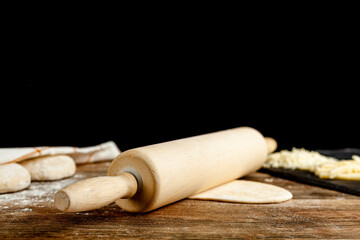 An American wooden rolling pin on  a flattened dough on wood kitchen countertop. A bread making concept image with loafs and cheese toppings in blurred background.